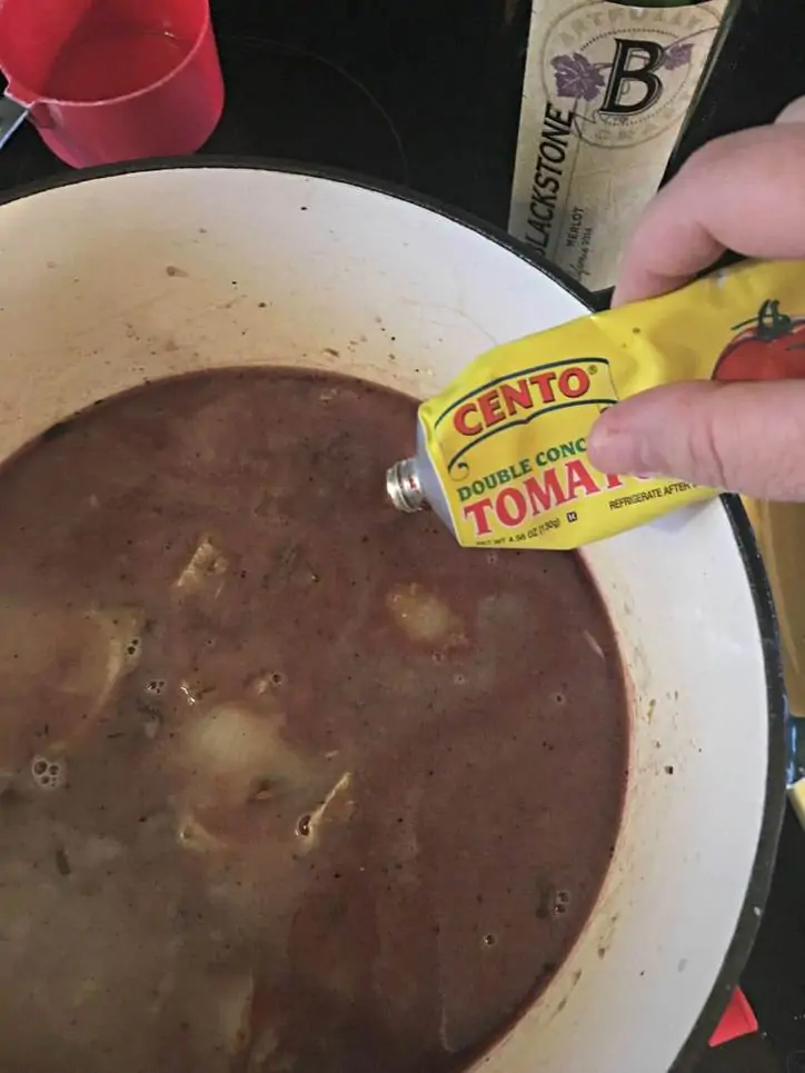 Tomato paste being squeezed into the broth in a duth oven with wine and measuring cup in background