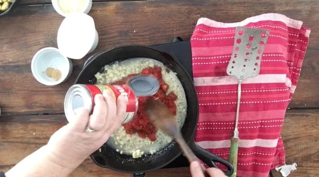 Adding a can of tomatoes to the pan that has garlic and shallot. Red towel and a spatula to the right