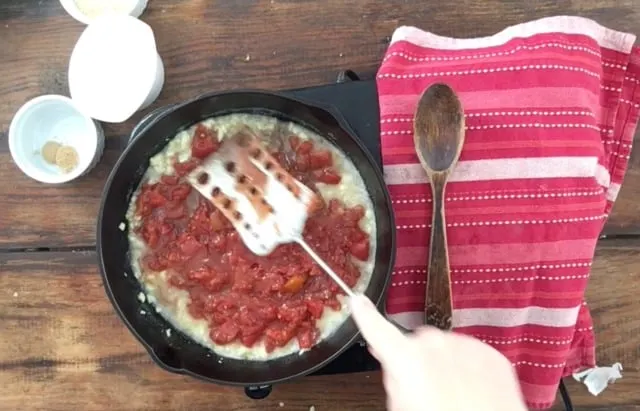 Smashing the tomatoes in the cast iron skillet with a spatula. Red towel and wooden spoon to the right.