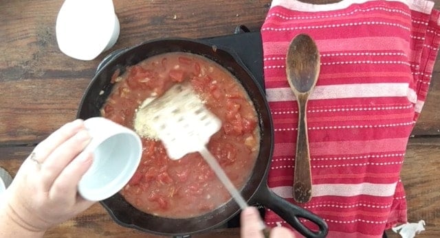 Adding the other ingredients to the cast iron skillet with red towel and wooden spoon to the right for southern tomato gravy