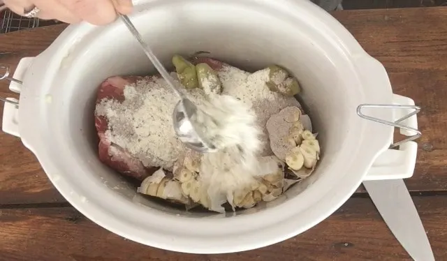 A tablespoon measure adding ranch seasoning to the crockpot with the chuck roast, garlic and other ingredients.