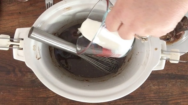 Pouring cornstarch slurry out of a glass measure to the drippings to make a gravy in the crockpot with whisk in the back ground FOR ALL THE SECRETS TO PERFECT CROCKPOT POT ROAST