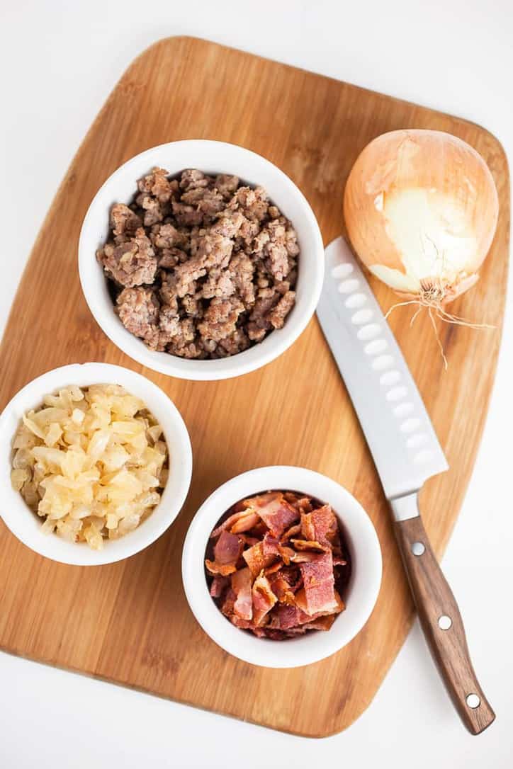 Small bowls of sausage, onions, bacon a knife, cutting board and whole onion