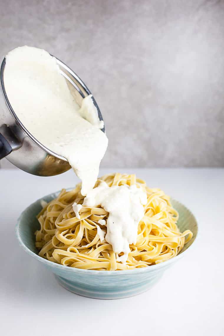 A photo of the a small pan of Alfredo sauce being poured over the fettuccini noodles in a blue bowl