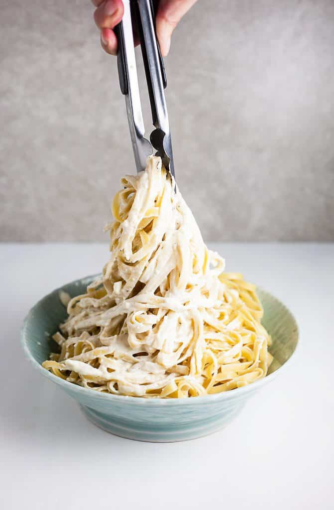 A photo of the noodles in a pair of tongs having the sauce worked in being swirled in a blue bowl