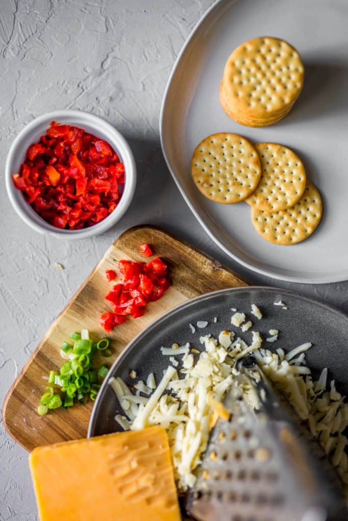 white plate with crackers, cutting board with cheese, pimentos and scallions
