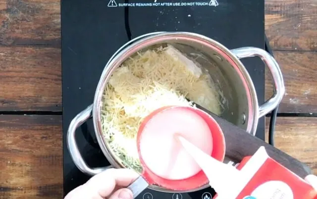 A measuring cup of cream over the top of the sauce pan with butter, cream and parmesan cheese in it.