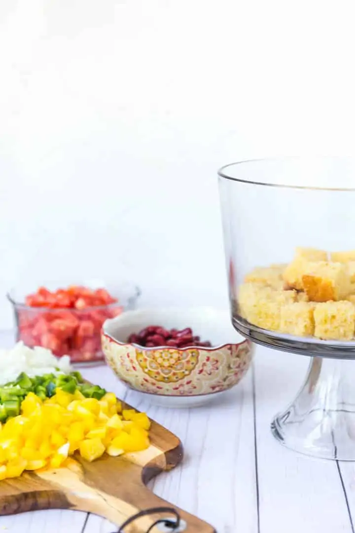 A photo of a glass trifle bowl with cornbread in the bottom, cutting board of ingredients.