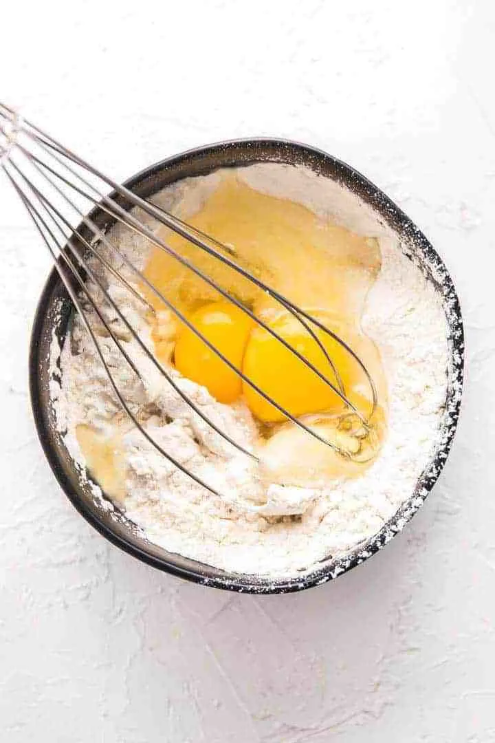 A photo of a black bowl, two eggs, flour and a whisk