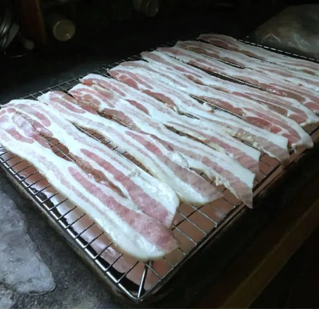 A photo of strips of raw bacon laying on top of the rack on top of a rimmed baking sheet