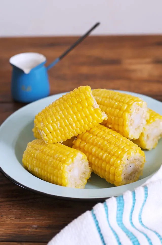 A photo of a blue plate of yellow corn on the cob with butter in a blue urn