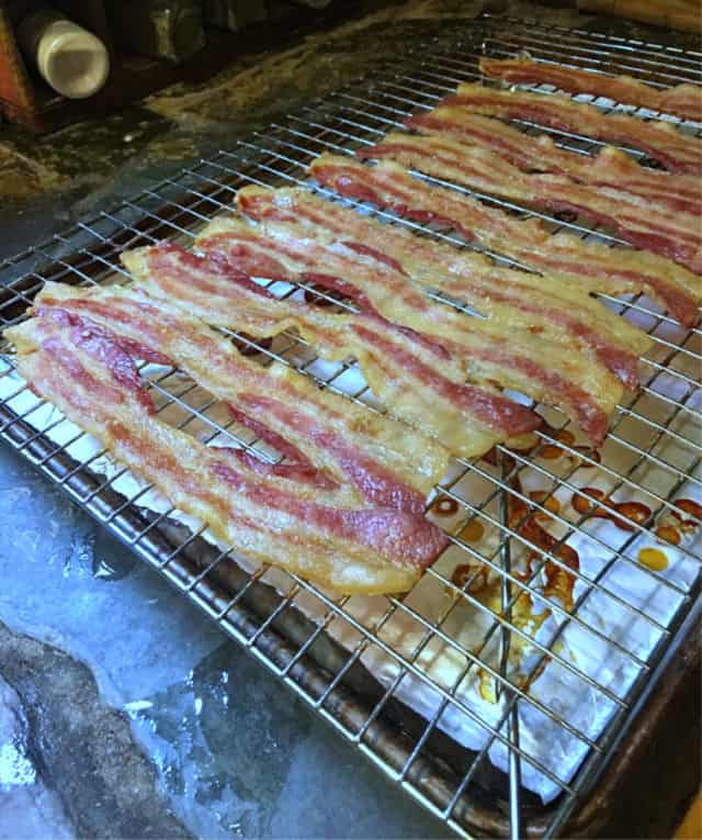 Stips of cooked bacon on a rack in a rimmed baking sheet. Drips of cooked grease on the foil in the bottom of the pan