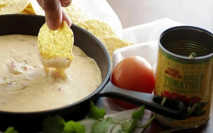 A photo of a chip dipping into the queso with cheese dripping off and a tomato