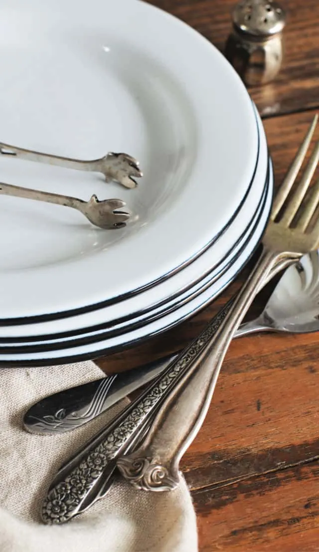 A stack of 4 white plates, silverware, pickle tongs and napkin