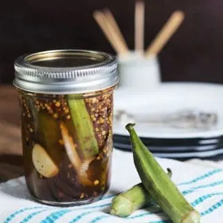 A jar of pickled okra and two okra pods sitting on a white and blue towel with plates and toothpicks in background