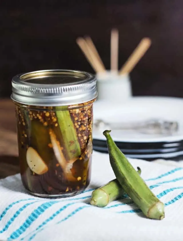 A jar of pickled okra and two okra pods sitting on a white and blue towel with plates and toothpicks in background