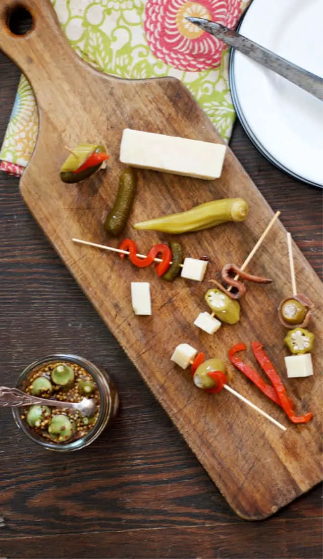 A cutting board of appetizers using pickled okra, cheese, pickles and red peppers