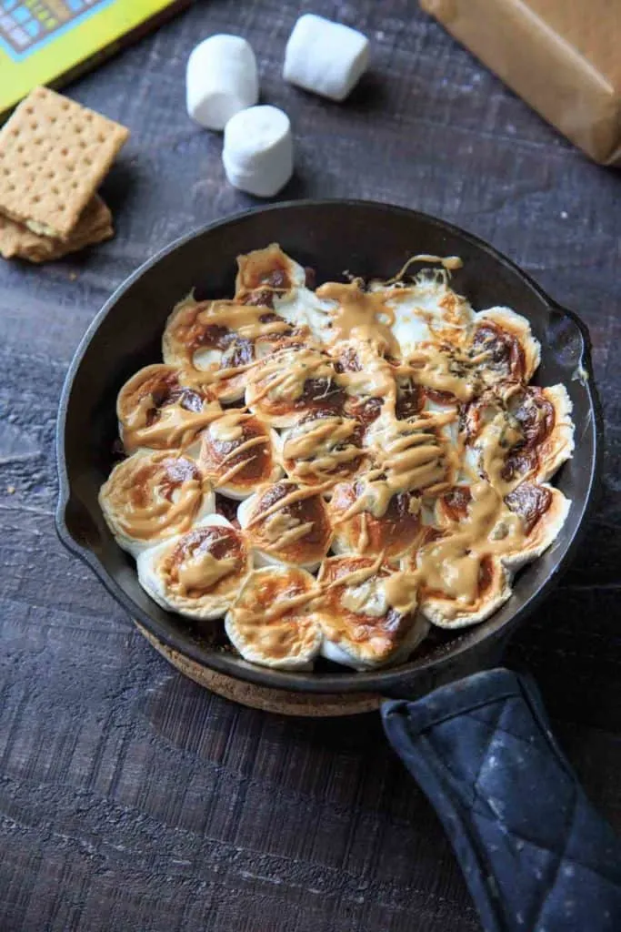 A cast iron skillet full of smores with marshmallows in the background and graham crackers