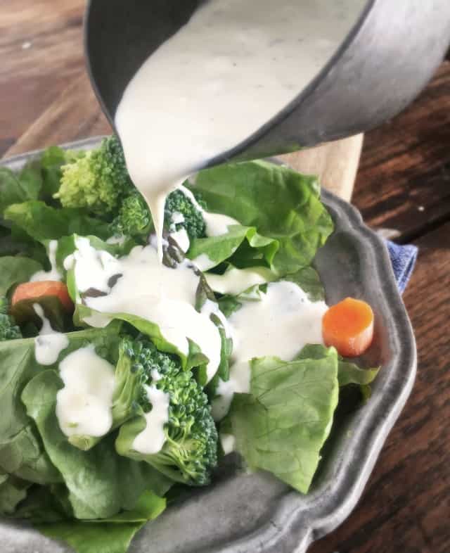 Silver bowl pouring restaurant ranch dressing onto green salad on grey plate