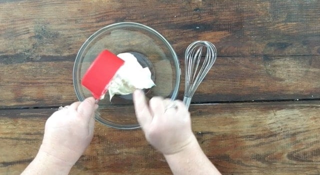 a photo of a cup measure with mayonnaise pouring into a glass bowl with a whisk to the right