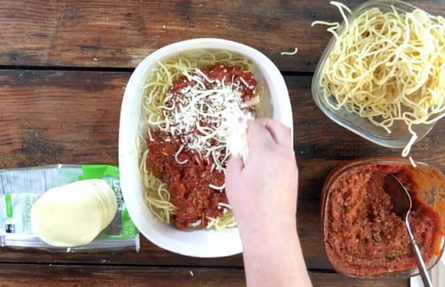 The first layer of spaghetti noodles, sauce and beginning to sprinkle the cheese in a white dish