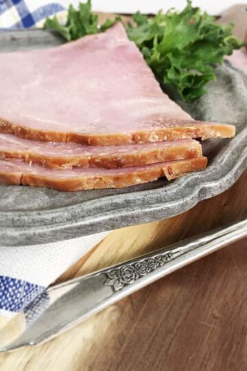 The edge of three pieces of ham on a gray plate with a napkin and the handle of a fork