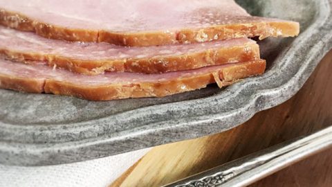 The edge of three pieces of ham on a gray plate with a napkin and the handle of a fork