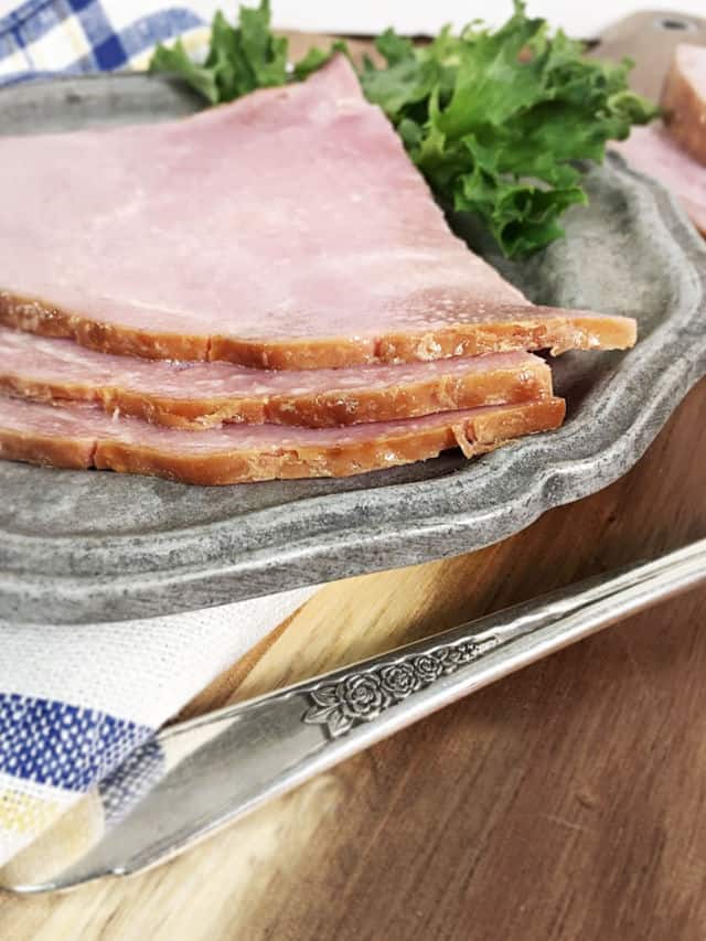 The edge of three pieces of ham on a gray plate with a napkin and the handle of a fork