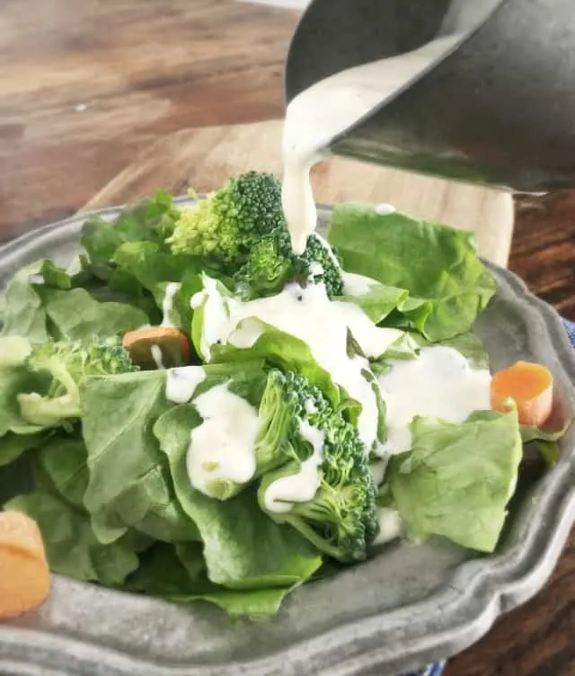 Creamy white restaurant ranch dressing pouring onto a green salad with orange carrots and green broccoli on a grey plate