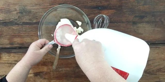 A photo of pouring milk into a cup measure for Restaurant Ranch Dressing. Large glass bowl and whisk to the right.