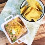 An overhead shot of a square bowl of refried beans with a small blue bowl of tortilla chips on a green napkin