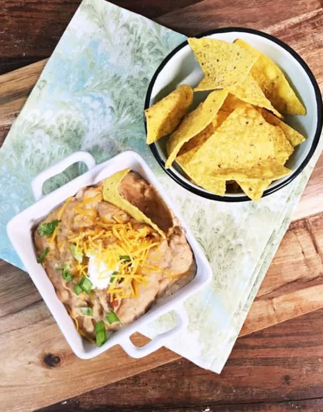 An overhead shot of a square bowl of refried beans with a small blue bowl of tortilla chips on a green napkin