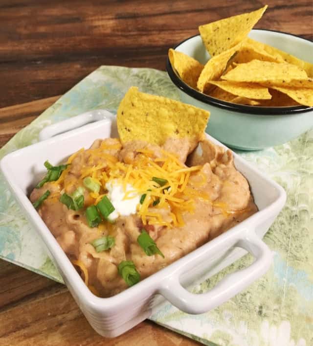 A square bowl of refried beans with sour cream, shredded cheddar and a chip stuck in the top