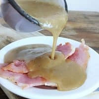 A blue cup of red eye gravy pouring over three pieces of country ham on a white plate