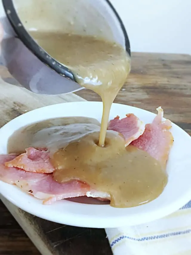 A blue cup of red eye gravy pouring over three pieces of country ham on a white plate