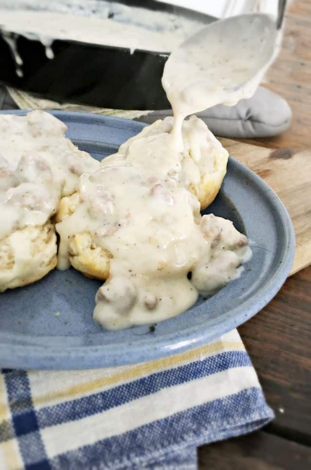 Sawmill Gravy being poured from a ladle onto biscuits sitting on a blue plate with black skillet in background