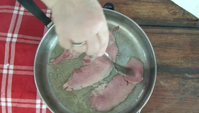 three slices of country ham frying in a skillet with red towel