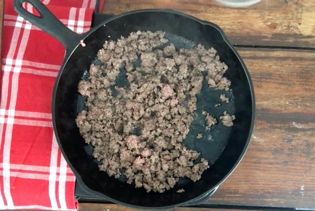 ground beef browning in a cast iron skillet with red checked towel to side