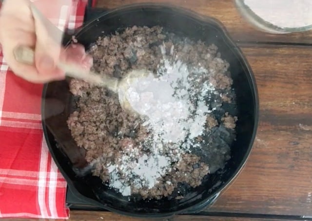 a photo of a spoon of flour sprinkling across the hamburger in the skillet with red towel at side. 