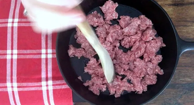 Wooden spoon chopping ground beef in a black iron skillet with red towel 