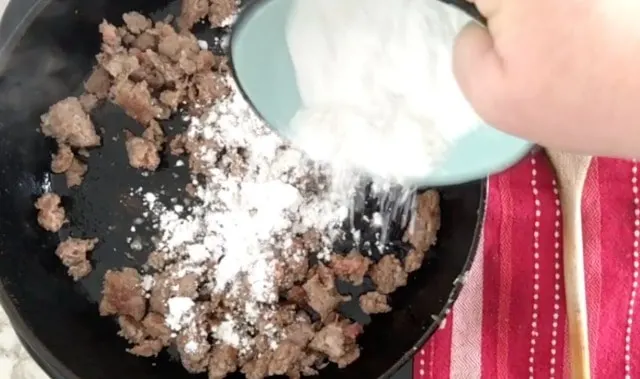 A photo of flour in a blue bowl being sprinkled over the sausage in the cast iron skillet