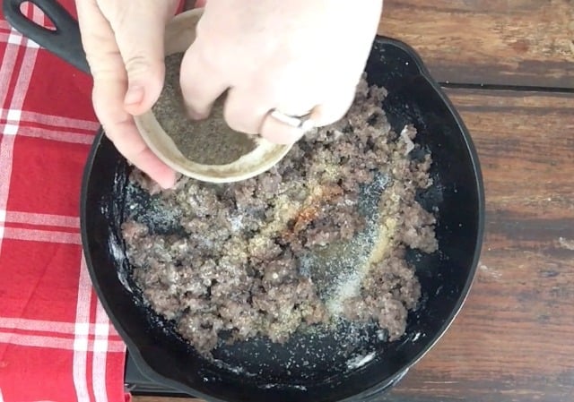 a photo of hands adding pepper to the skillet of browned ground beef with other seasonings in it and red towel to the side