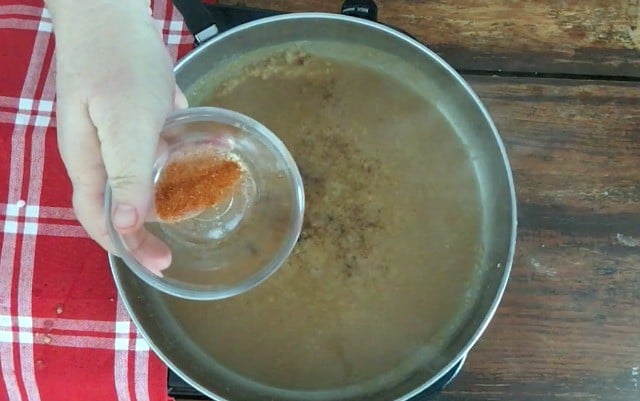 A small bowl of cayenne pepper held over the top of a silver skillet with gravy in it
