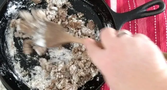 a photo of the sausage in the cast iron skillet with flour on it being stirred and cooked using a wooden spoon