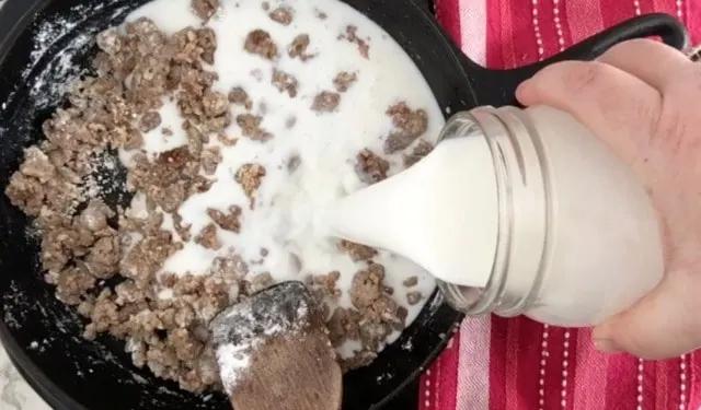 A photo of a quart jar pouring milk into a black cast iron pan with sausage and flour in it