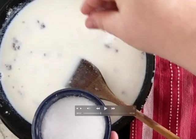 a photo of salt in a small blue bowl being added to the cast iron pan mixture with wooden spoon in photo