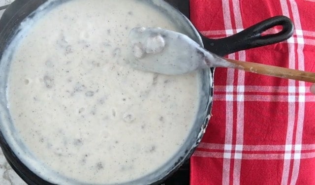 a photo of a wooden spoon covered with gravy and clumps of sausage over the top of a cast iron skillet filled with milky, creamy thick gravy