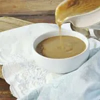 A gravy boat pouring gravy into a small white bowl sitting on a white napkin with wood table