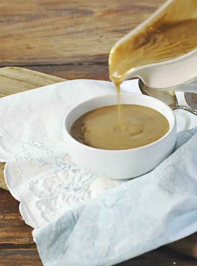 A gravy boat pouring gravy into a small white bowl sitting on a white napkin with wood table