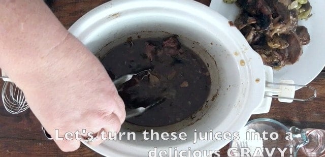 a photo of hand with tongs removing bits of meat from a crockpot leaving the juices behind. Plate of meat in back ground